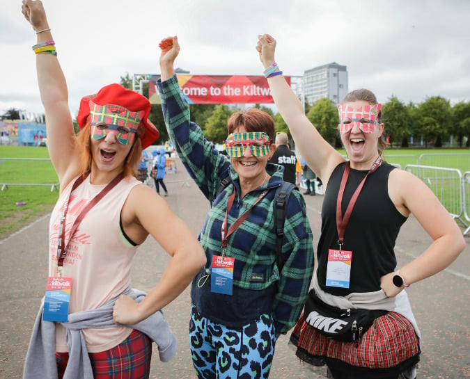 Three woman at the Kiltwalk start line standing together with one hand in the air smiling at the camera