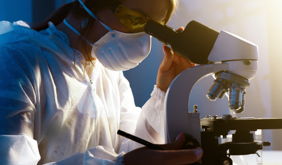 woman looking through a microscope with a mask and goggles on