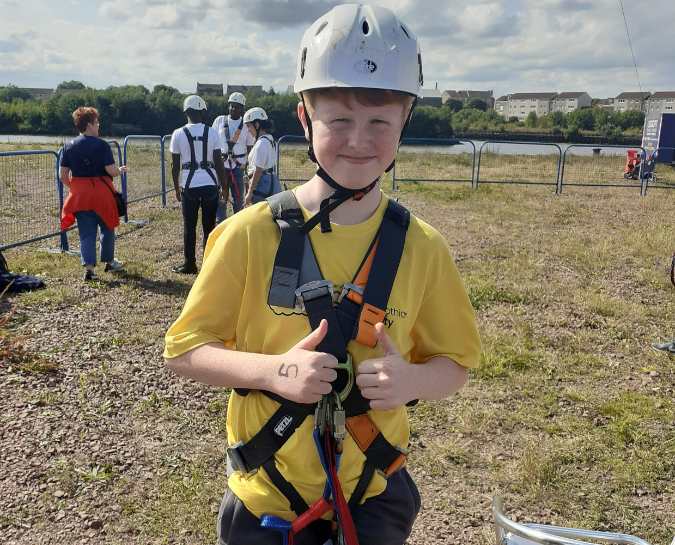 NHS Lothian Charity Champion smiling at the camera with their thumbs up. Getting ready for their zipslide with their harness and helmet on and a yellow Fighting Against Cancer Edinburgh t-shirt on.