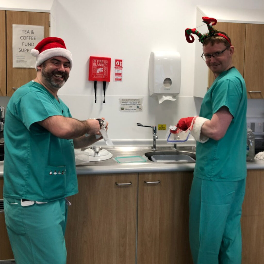 2 members of NHS Lothian staff wearing a Christmas hat and reindeer antlers smiling at the camera whilst washing up on the RHCYP PCCU ward.