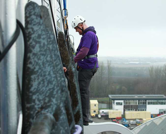 Side view of a NHS Lothian Charity Champion starting the decent from the Falkirk Wheel Abseil. He is stood up at the edge waiting to start the decent. Wearing a harness, helmet and a purple NHS Lothian Charity t-shirt.