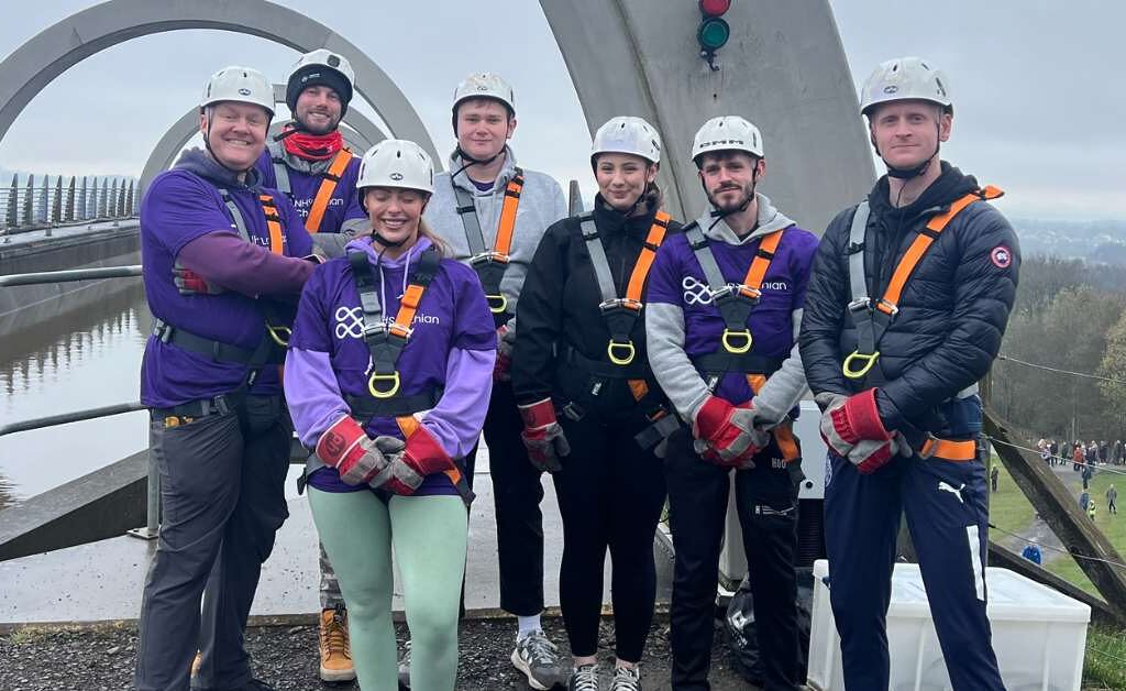 Seven NHS Lothian Charity Champions stood smiling at the camera waiting for their turn to do the abseil. Wearing a harness, helmet and a purple NHS Lothian Charity t-shirt.