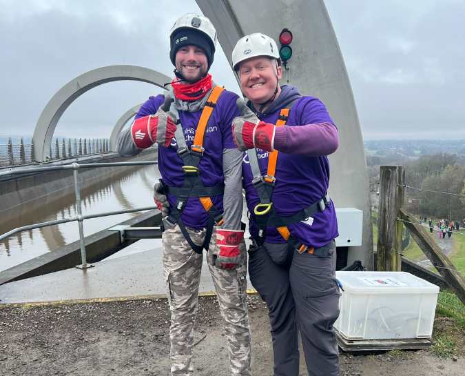Two NHS Lothian Charity Champions stood smiling at the camera with their thumbs up waiting for their turn to do the abseil. Wearing a harness, helmet and a purple NHS Lothian Charity t-shirt.