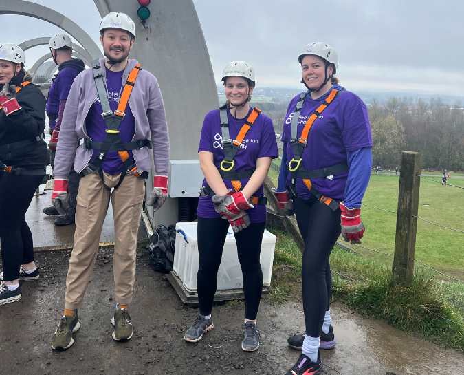 Three NHS Lothian Charity Champions stood smiling at the camera waiting for their turn to do the abseil. Wearing a harness, helmet and a purple NHS Lothian Charity t-shirt.