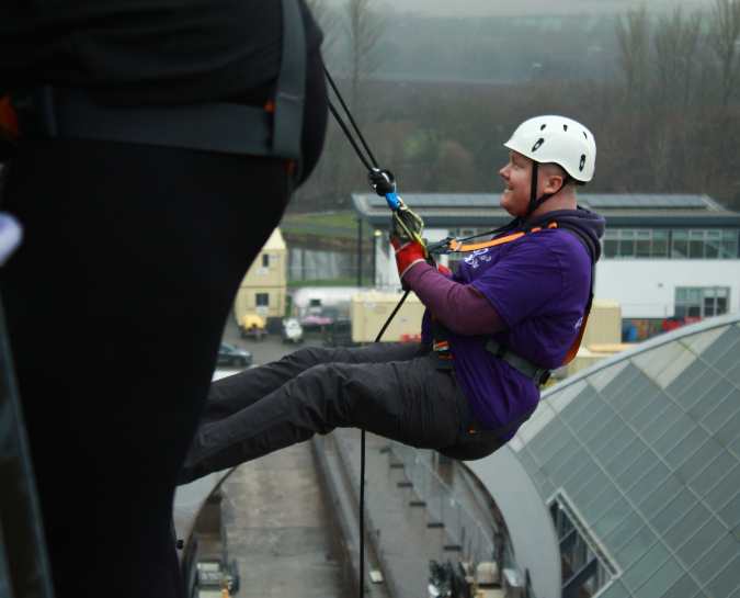 Side view of a NHS Lothian Charity Champion starting the decent from the Falkirk Wheel Abseil. Wearing a harness, helmet and a purple NHS Lothian Charity t-shirt.