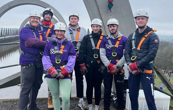 Seven NHS Lothian Charity Champions stood smiling at the camera waiting for their turn to do the abseil. Wearing a harness, helmet and a purple NHS Lothian Charity t-shirt.