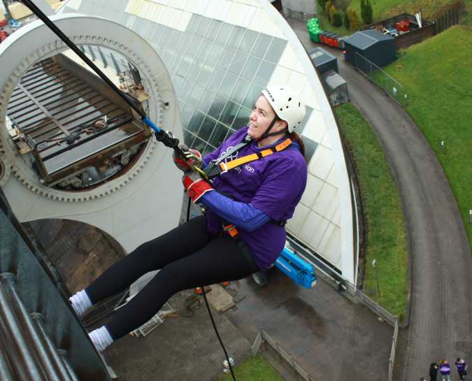 Side view of a NHS Lothian Charity Champion starting the decent from the Falkirk Wheel Abseil. Wearing a harness, helmet and a purple NHS Lothian Charity t-shirt