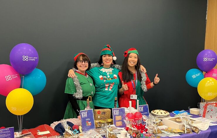 Three woman smiling at the camera dressed as elves. They are stood behind a charity table selling cakes to support NHS Lothian Charity