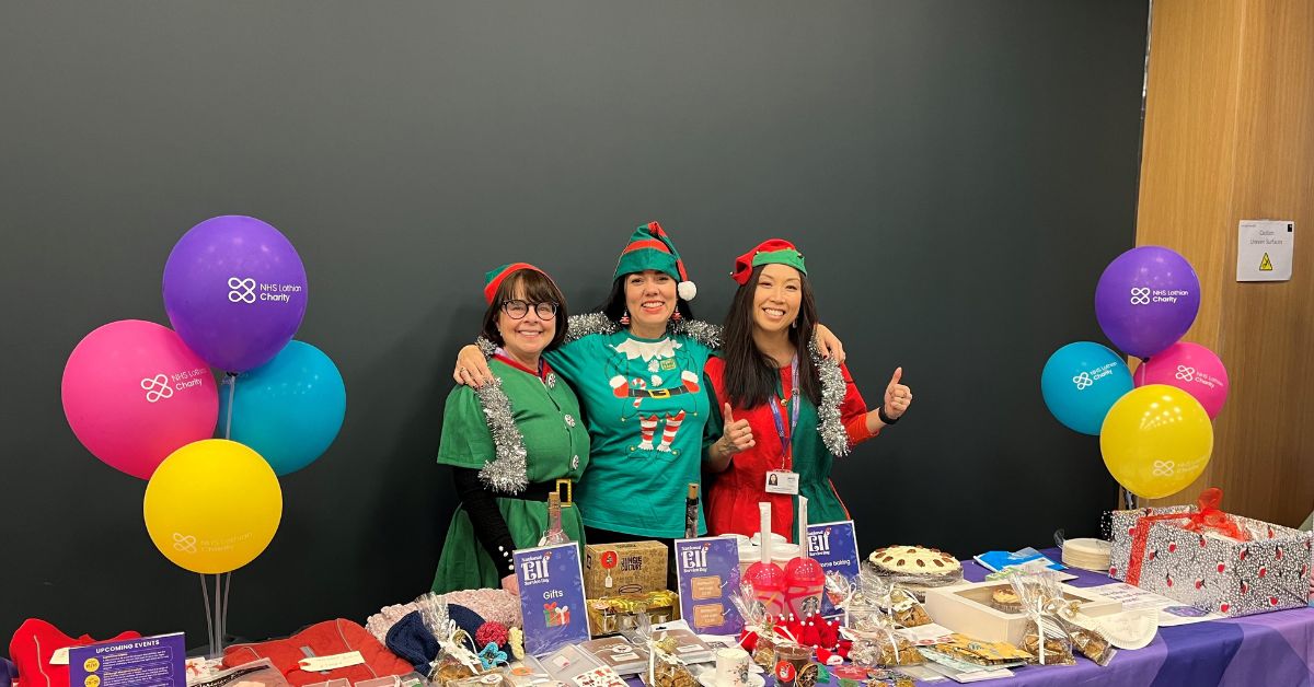 Three woman smiling at the camera dressed as elves. They are stood behind a charity table selling cakes to support NHS Lothian Charity