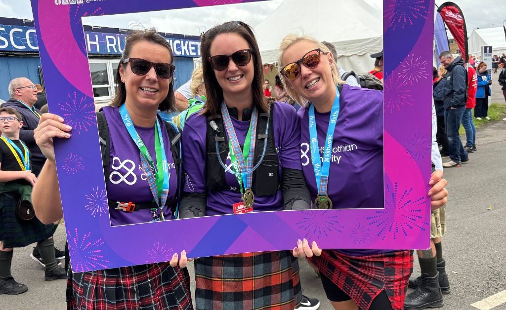 three kiltwalksers with a finishers board dressed in tartan at the end of the Kiltwalk