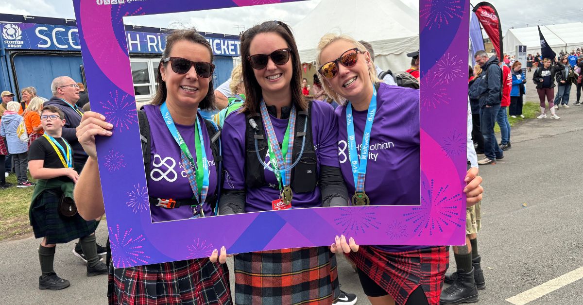 three kiltwalksers with a finishers board dressed in tartan at the end of the Kiltwalk