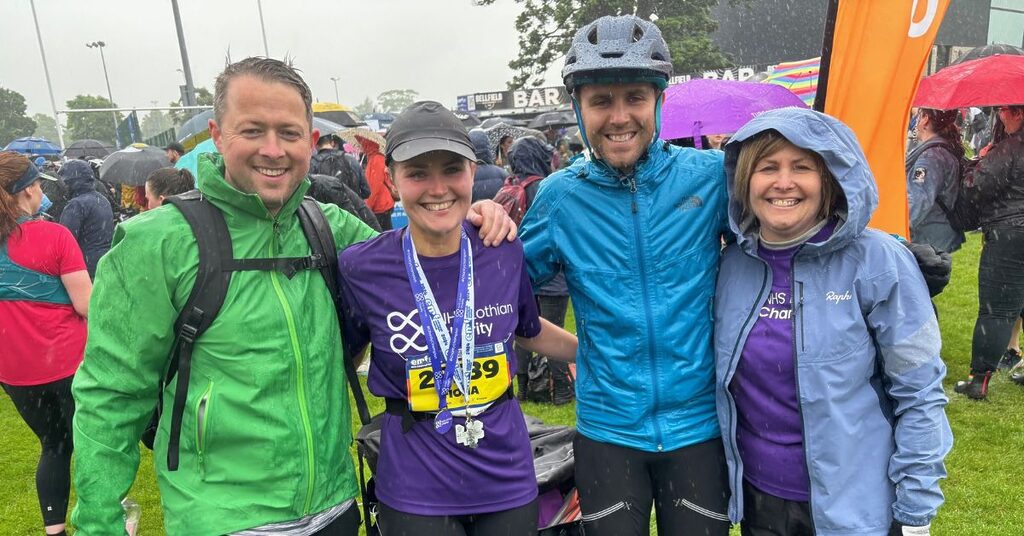 four people standing in the rain in a field after the Edinburgh Marathon Festival with a crowd of people behind them