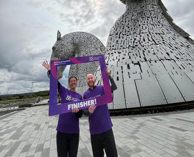 Two NHS Lothian Charity fundraisers holding a finisher frame in front of the Kelpies horses