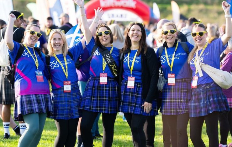 Group of Ladies smiling wearing NHS Lothian Charity t-shirts and kilts at the Kiltwalk starting line